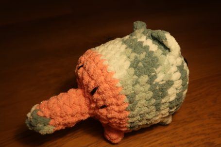 A small, crocheted toy with a plump body and a long, rounded snout, in pastel colours.